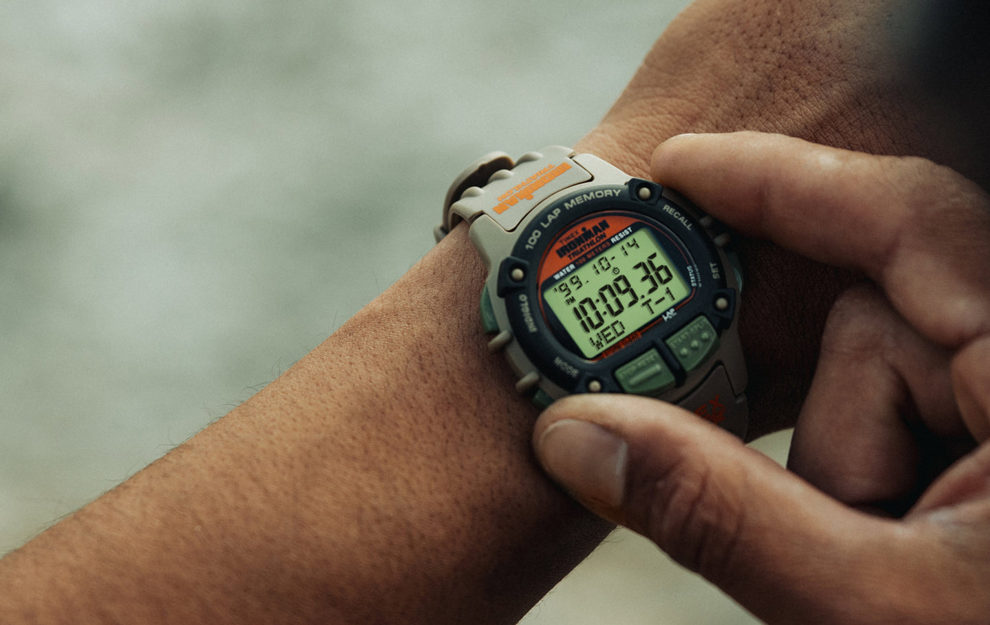 A close-up of a person wearing a TIMEX® IRONMAN® Flix x Huckberry digital sports watch on their wrist. The watch has a rugged gray and black case with green buttons and an orange logo. Its digital display shows the time 10:09:36, the date Wednesday 10-14, and various stopwatch and lap memory indicators.