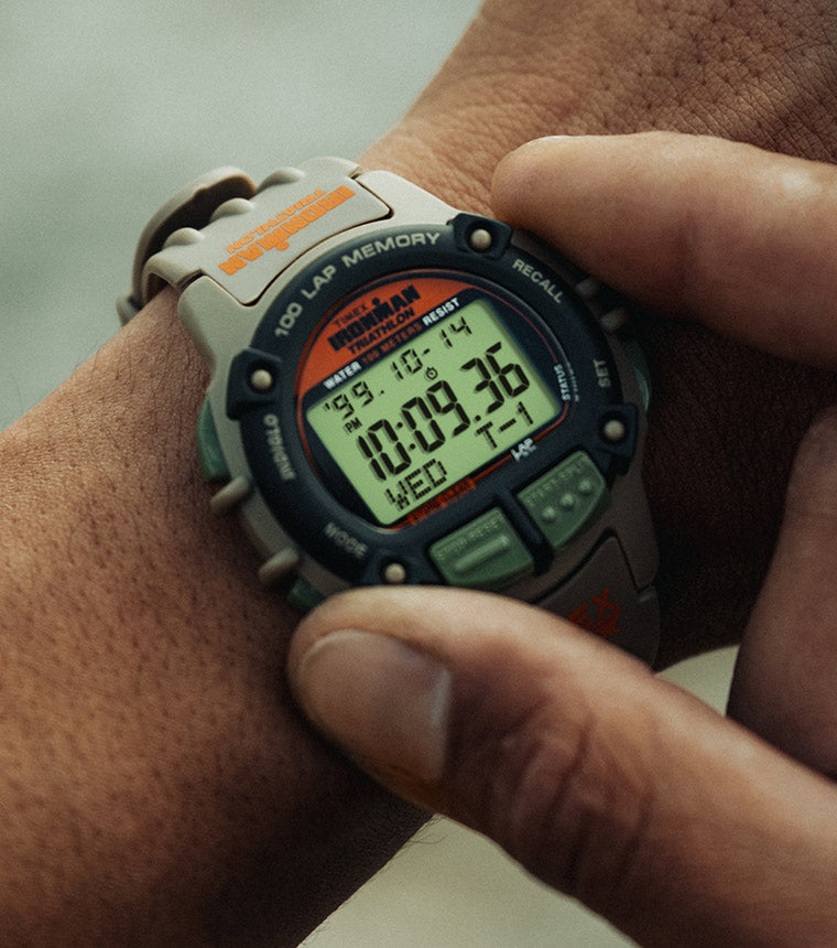 A close-up of a person wearing a TIMEX® IRONMAN® Flix x Huckberry digital sports watch on their wrist. The watch has a rugged gray and black case with green buttons and an orange logo. Its digital display shows the time 10:09:36, the date Wednesday 10-14, and various stopwatch and lap memory indicators.