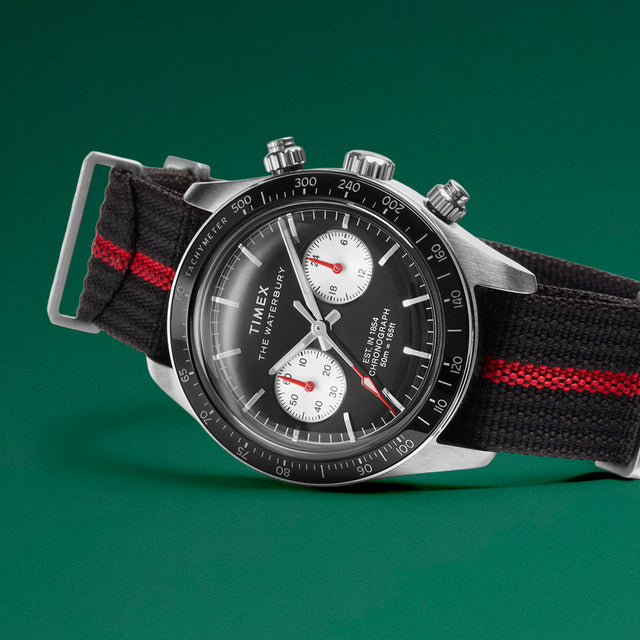 Close-up of the Timex Waterbury Heritage Chronograph resting on a green surface, featuring a brushed stainless-steel case, tachymeter bezel, and dual subdials.