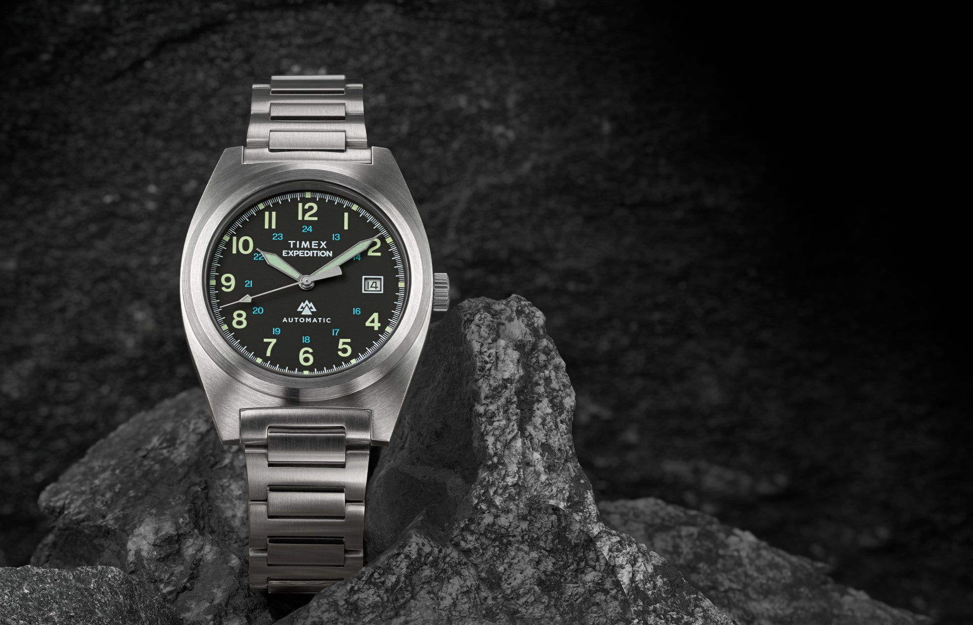 A Timex Expedition® Capstone Automatic watch with a brushed stainless-steel case and bracelet rests among rugged gray rocks. The black dial features luminous green Arabic numerals, a date window at 3 o’clock, and bold hands designed for outdoor durability.