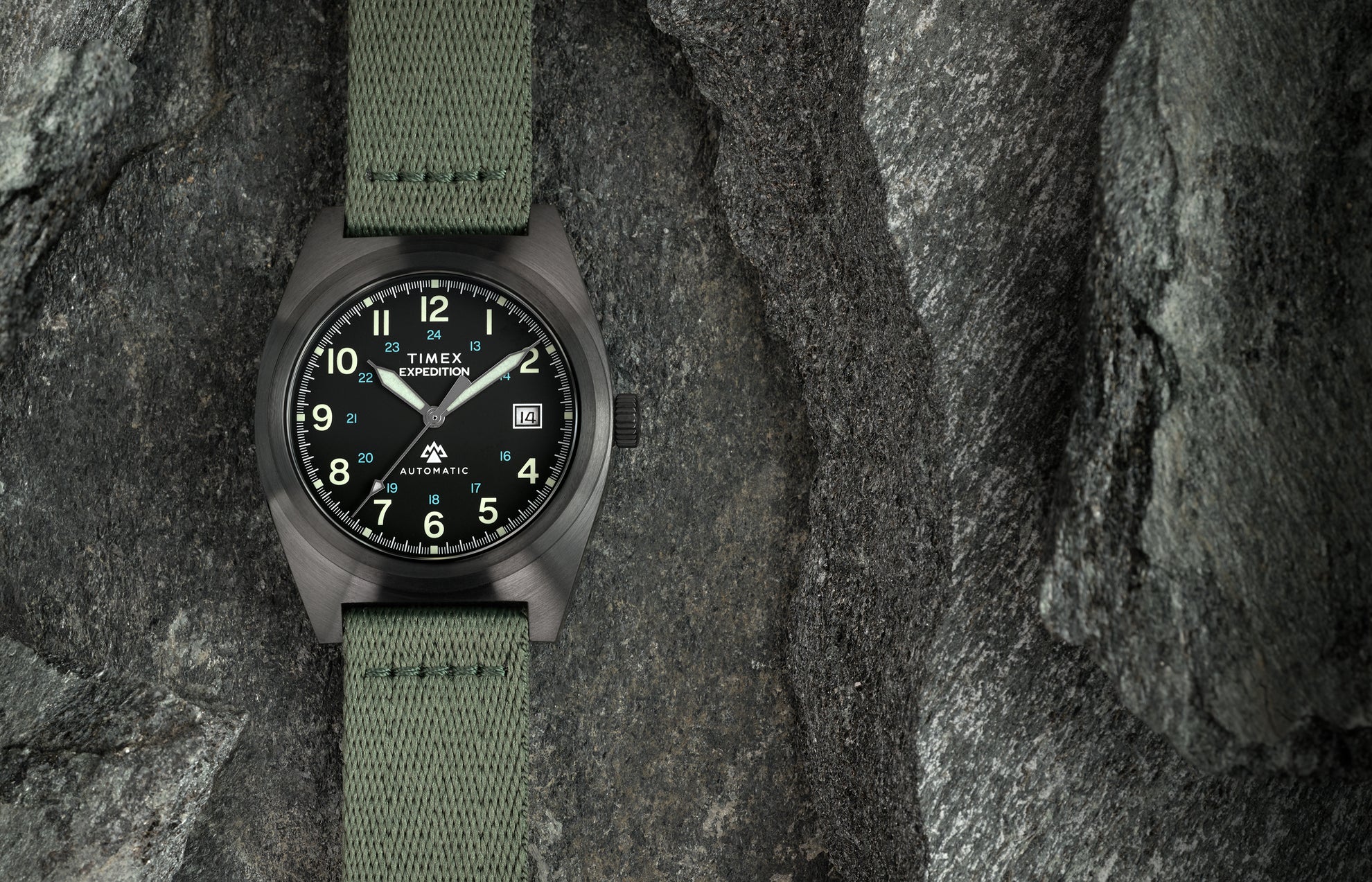 A Timex Expedition® Capstone Automatic watch with a matte gunmetal case and green fabric strap rests among rugged stone surfaces. The black dial features luminous green numerals, blue 24-hour markings, and a date window at 3 o’clock, emphasizing its field-ready durability.