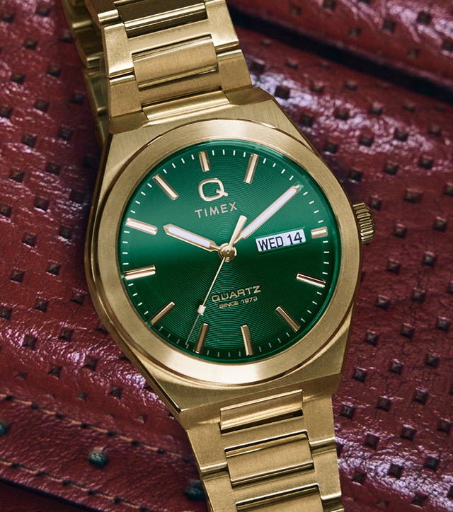 A gold-tone Q Timex® Continental Day and Date watch with a vibrant green dial rests on a red perforated leather car seat. The watch features a brushed stainless-steel bracelet, polished bezel, and a day-date display at 3 o’clock, combining bold color with vintage-inspired design.