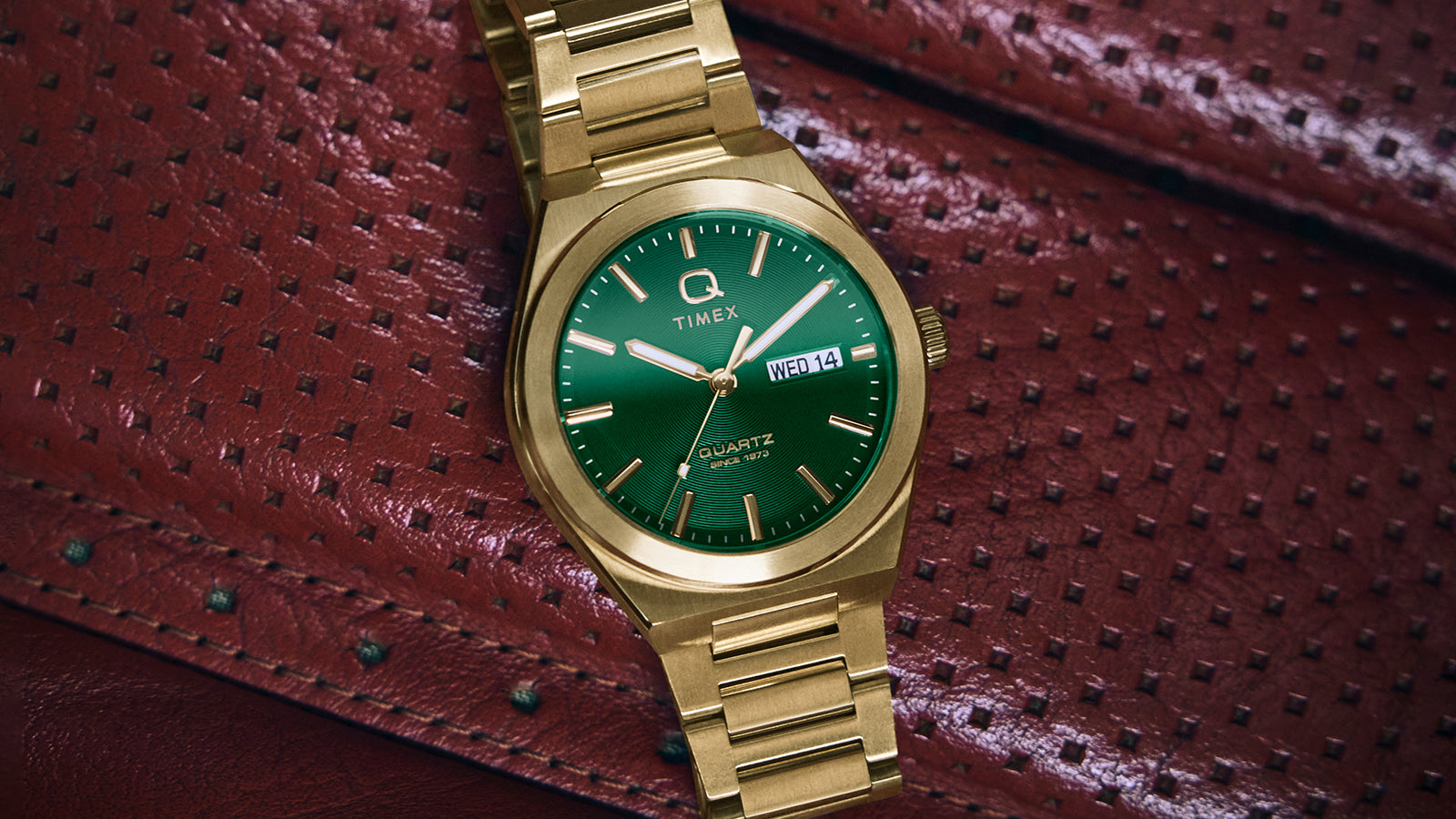 A gold-tone Q Timex® Continental Day and Date watch with a vibrant green dial rests on a red perforated leather car seat. The watch features a brushed stainless-steel bracelet, polished bezel, and a day-date display at 3 o’clock, combining bold color with vintage-inspired design.