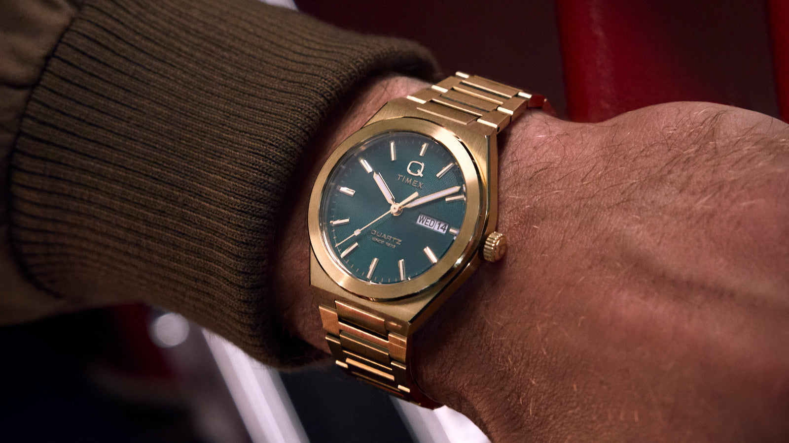 Close-up of a person wearing the gold-tone Q Timex® Continental Day and Date watch with a green dial. The elegant metallic finish and dark green face pop against the casual brown sleeve, showcasing a fusion of sophistication and everyday wearability.