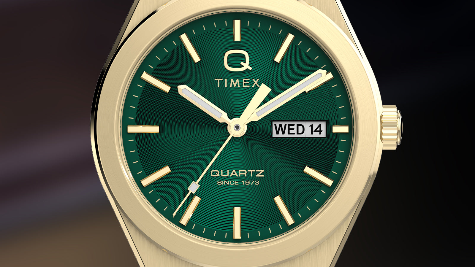 Macro shot of the gold-tone Q Timex® Continental Day and Date watch face, highlighting the textured green dial, luminous hands, and the “Q Timex” logo. The day-date window and gold-tone hour markers showcase precision craftsmanship and vintage styling.