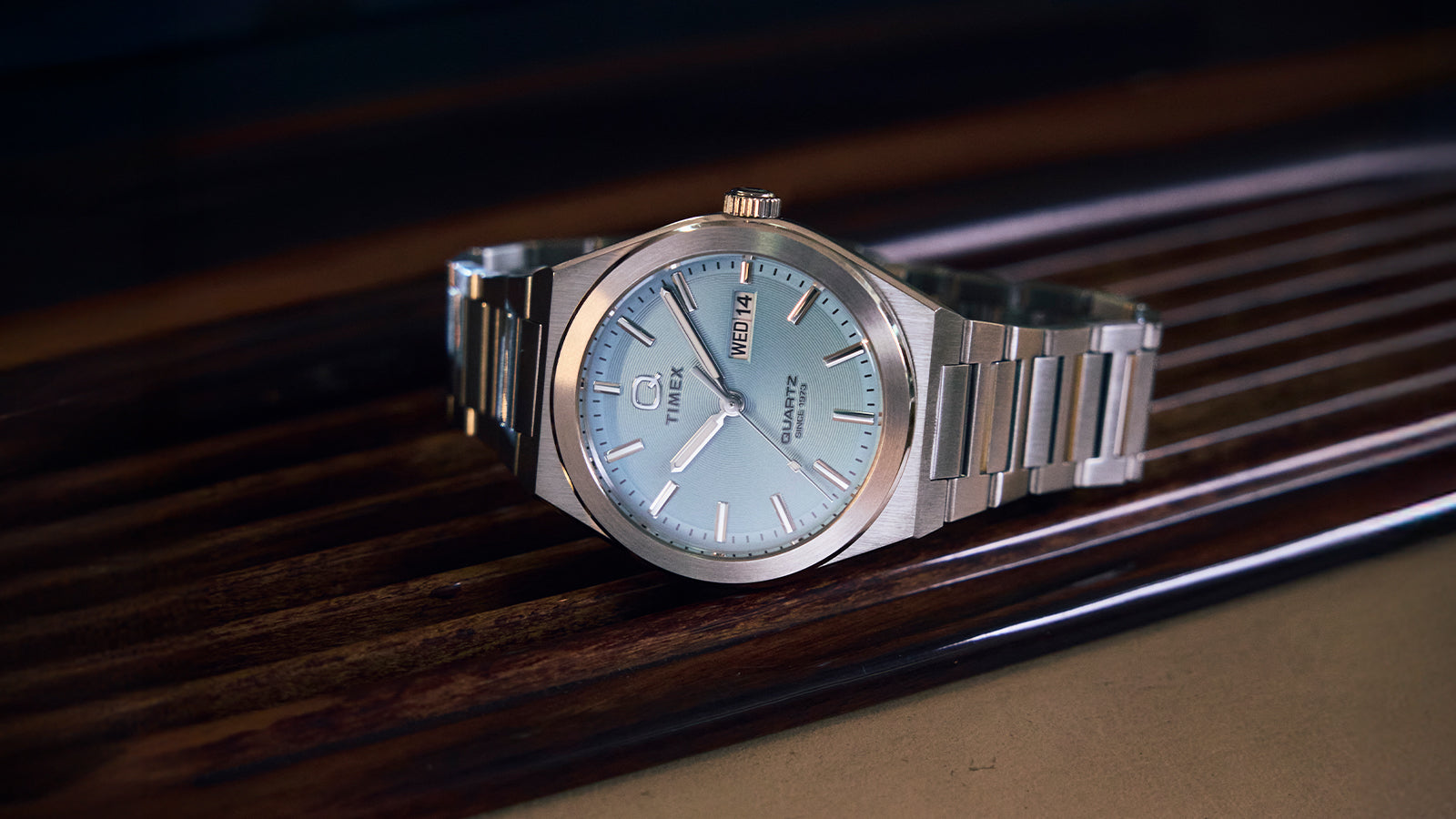 A stainless-steel Q Timex® Continental Day and Date watch with an ice-blue dial rests on a polished wooden car dashboard. The brushed metal case and bracelet emphasize its sleek, vintage-inspired design with a day-date window at 3 o’clock.