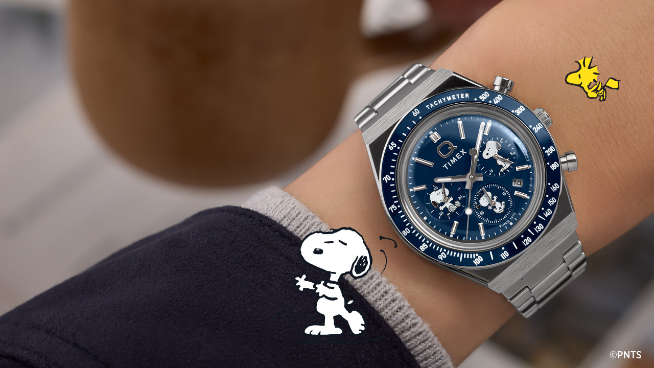 Person wearing the Timex x Peanuts® Q Timex® Chronograph featuring Snoopy and Woodstock on a blue dial with stainless steel bracelet. The playful Peanuts® illustrations complement the watch’s refined, retro-inspired chronograph design.