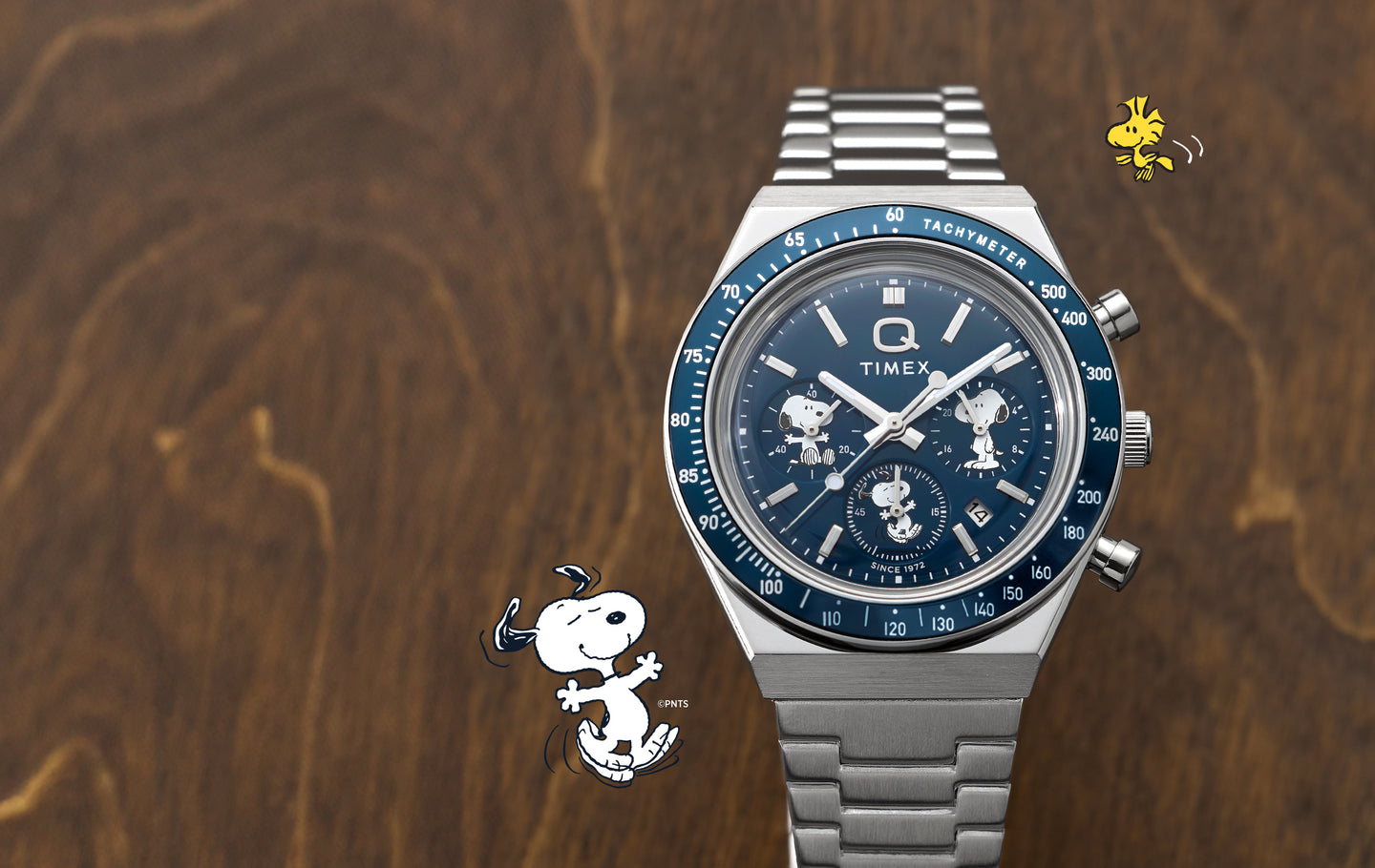 Timex Q Chronograph watch with a stainless-steel bracelet is shown on a wooden surface. The blue watch dial features Peanuts characters in the sub-dials, including Snoopy in different poses. Small illustrations of Snoopy dancing and Woodstock flying appear beside the watch, emphasizing the Peanuts collaboration.