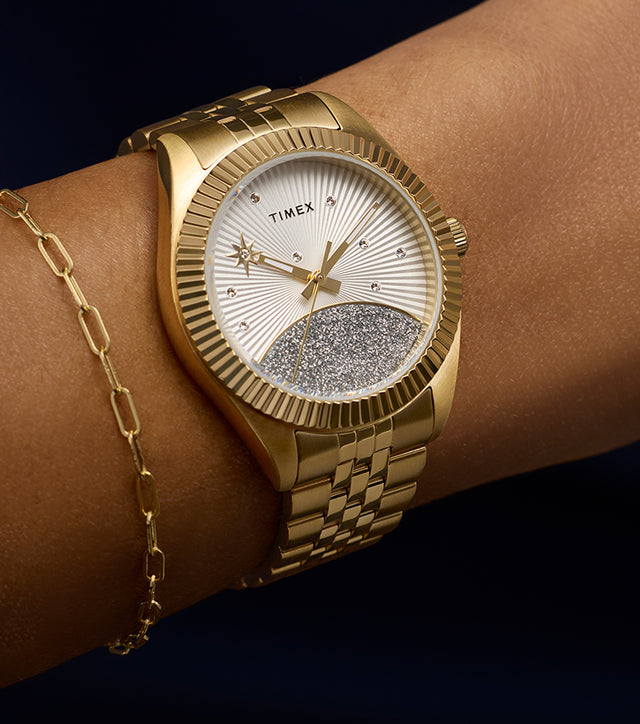 Timex Legacy watch in gold-tone stainless steel with a silver sunray dial featuring crystal accents and a glitter-textured lower half, worn on wrist with a delicate gold bracelet.