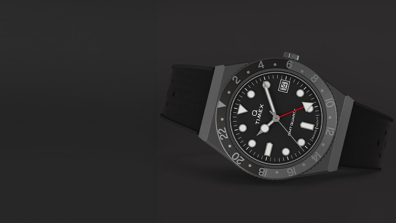 A Q Timex GMT watch with a black dial, red GMT hand, and black rotating 24-hour bezel resting on a dark surface, showcasing its sleek and functional aesthetic.