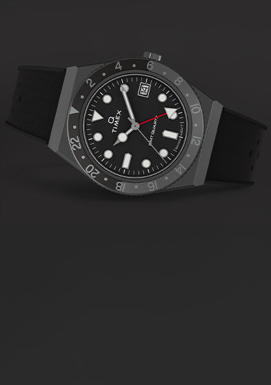 A Q Timex GMT watch with a black dial, red GMT hand, and black rotating 24-hour bezel resting on a dark surface, showcasing its sleek and functional aesthetic.