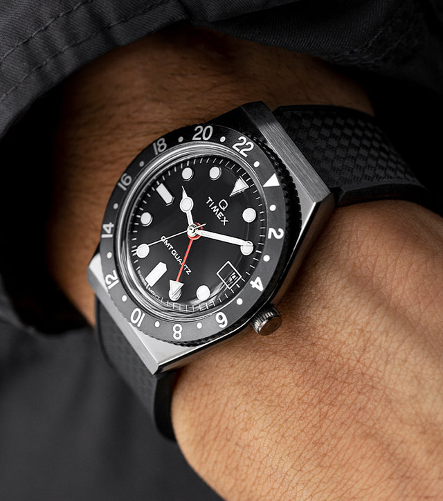 A Q Timex GMT watch with a black dial and red GMT hand worn on the wrist, styled with a black textured strap and matching dark jacket sleeve, highlighting its modern and rugged design.