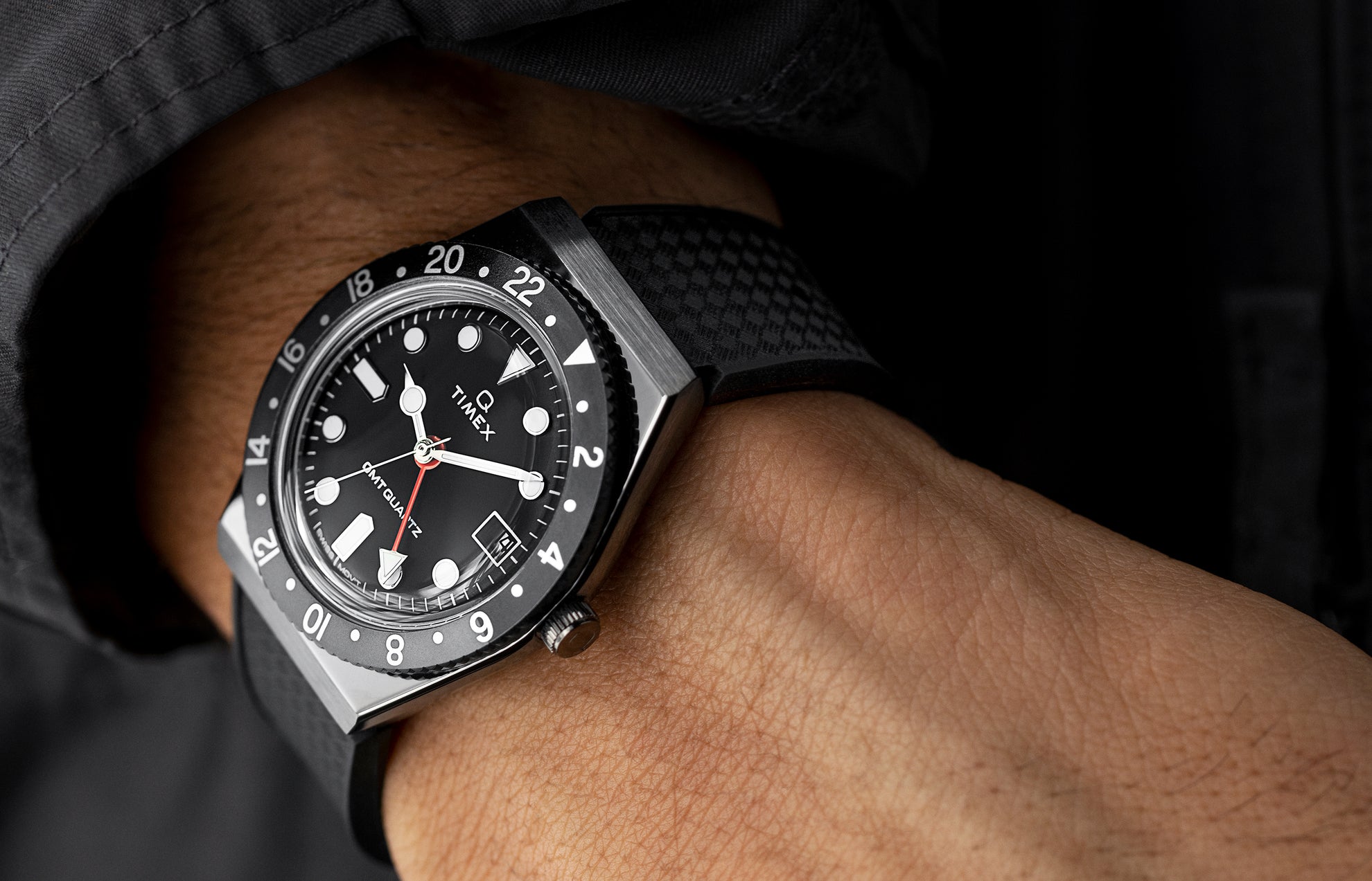 A Q Timex GMT watch with a black dial and red GMT hand worn on the wrist, styled with a black textured strap and matching dark jacket sleeve, highlighting its modern and rugged design.