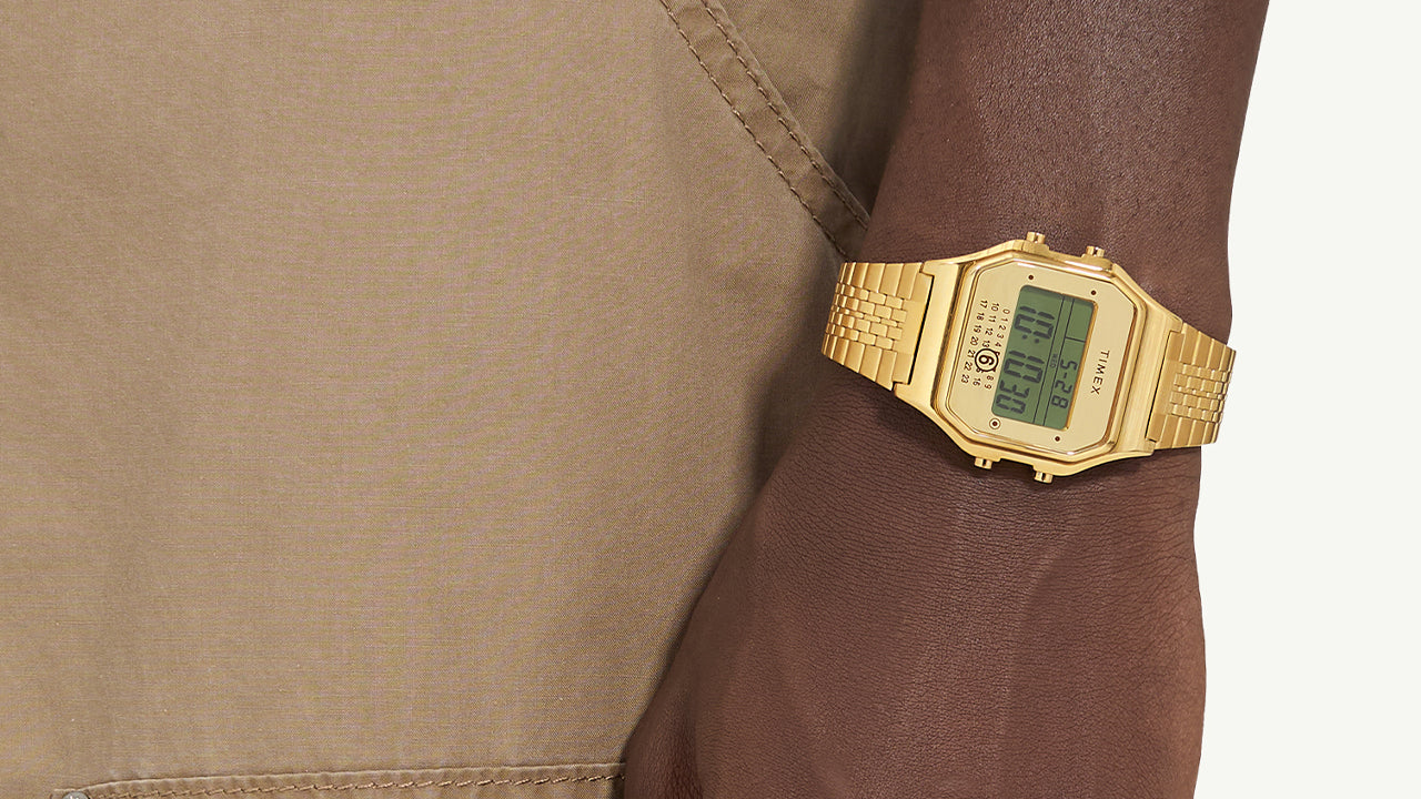 The gold Timex x MM6 T80 wristwatch worn on a darker skin tone model’s wrist, paired with tan pants, showing contrast and elegant wrist presence.
