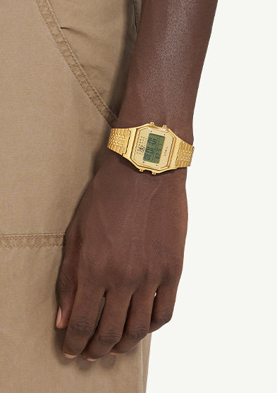 The gold Timex x MM6 T80 wristwatch worn on a darker skin tone model’s wrist, paired with tan pants, showing contrast and elegant wrist presence.