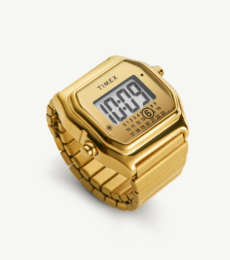 The Timex x MM6 Maison Margiela T80 digital watch in full gold-tone finish, featuring a square retro case, matching metal bracelet, and digital display. The minimalist dial includes the MM6 calendar-style date motif and Timex branding, set against a clean cream background.