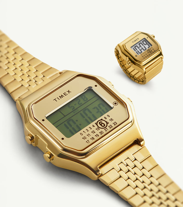 A close-up of two gold-tone digital timepieces from the Timex x MM6 Maison Margiela collection, featuring the signature Timex T80 digital watch and matching miniature ring watch. Both pieces display a retro digital screen with date and time on a sleek gold metal bracelet and ring band, set against a light background.