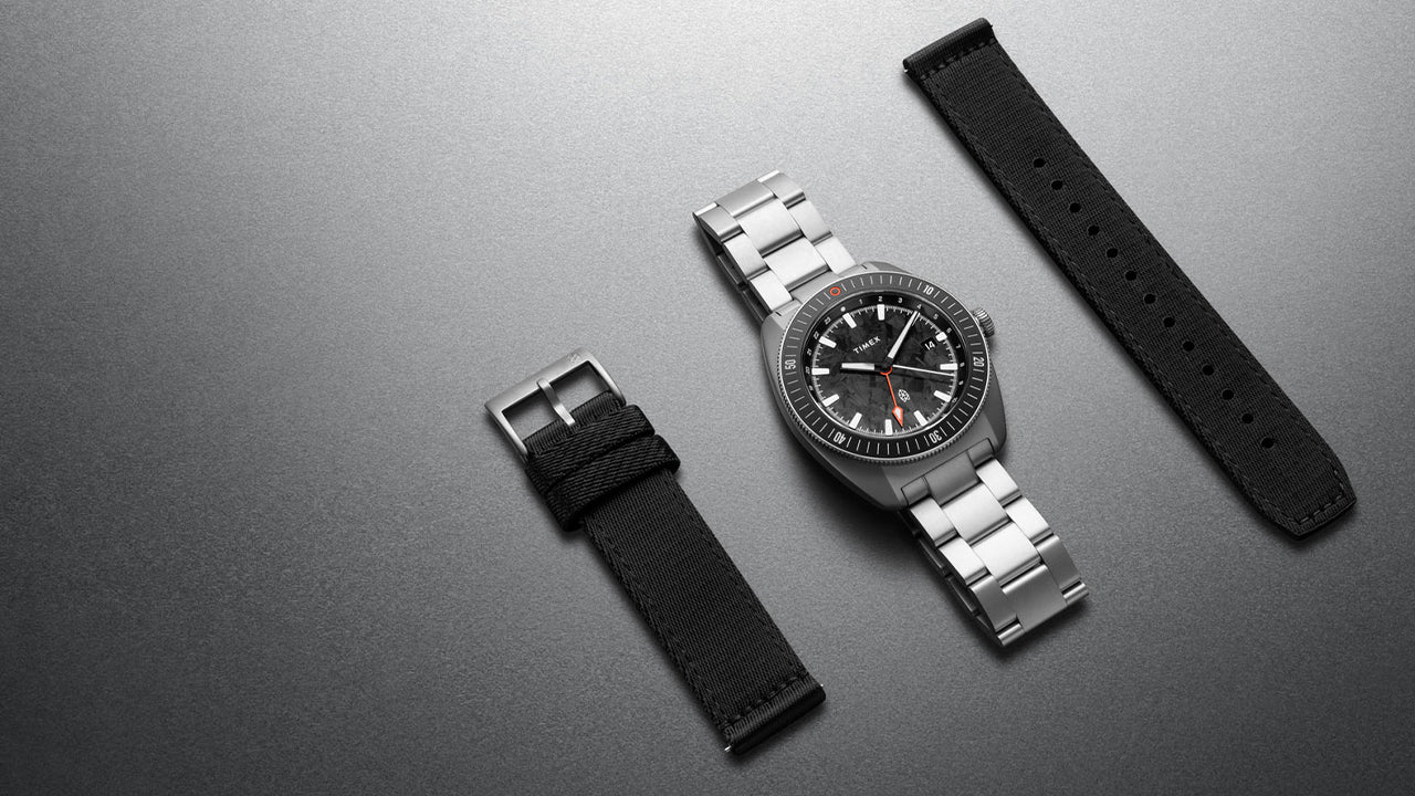 The James Brand x Timex GMT watch with stainless steel bracelet and interchangeable black fabric strap displayed on a gray surface.
