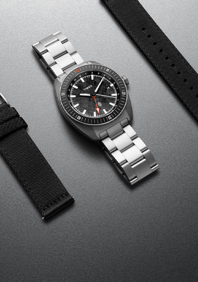 The James Brand x Timex GMT watch with stainless steel bracelet and interchangeable black fabric strap displayed on a gray surface.