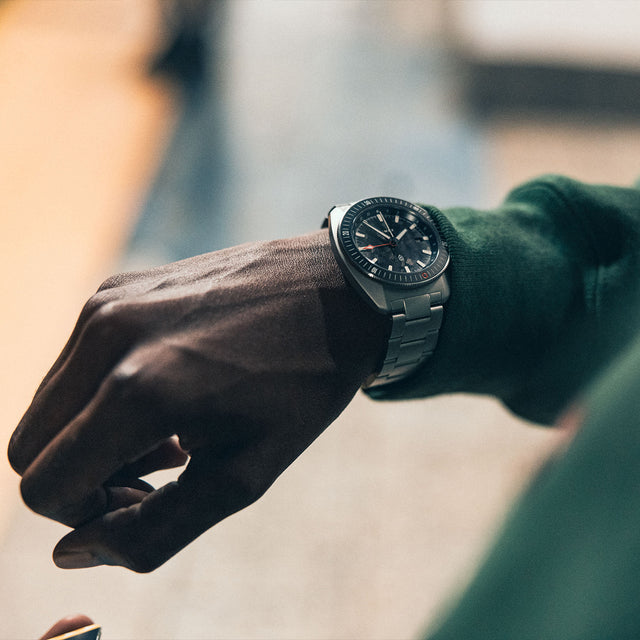 The James Brand x Timex GMT watch worn on the wrist, styled with a green sweatshirt, showing its sleek stainless steel design.