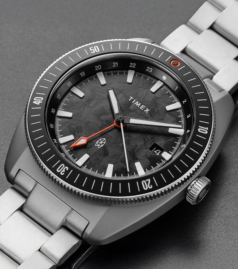 A close-up of The James Brand x Timex Automatic GMT watch featuring a black textured dial, orange GMT hand, brushed stainless steel case, and bracelet, displayed on a gray surface highlighting its modern and rugged aesthetic.
