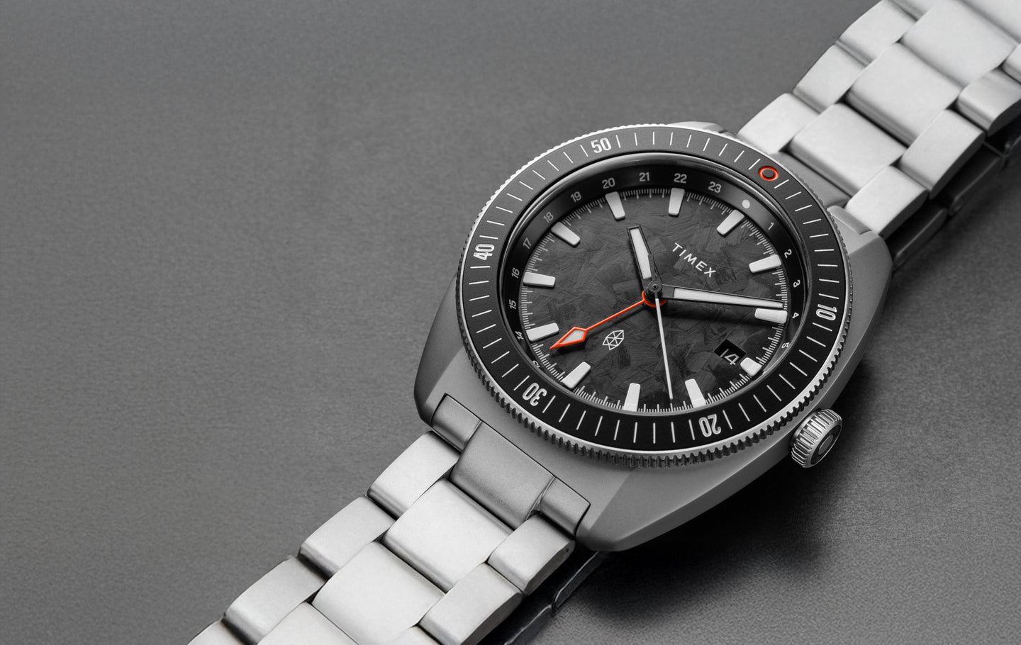 A close-up of The James Brand x Timex Automatic GMT watch featuring a black textured dial, orange GMT hand, brushed stainless steel case, and bracelet, displayed on a gray surface highlighting its modern and rugged aesthetic.