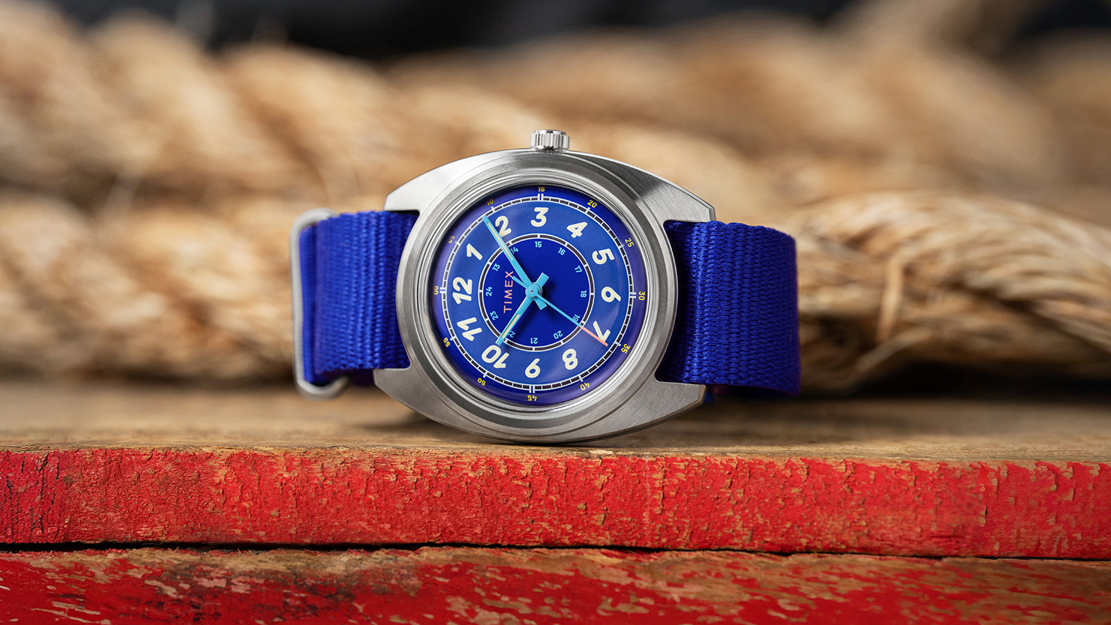 A Timex x Worn & Wound Whirlpool watch with a bright blue dial and matching blue fabric strap resting on a distressed red wooden surface, with thick coiled rope blurred in the background.