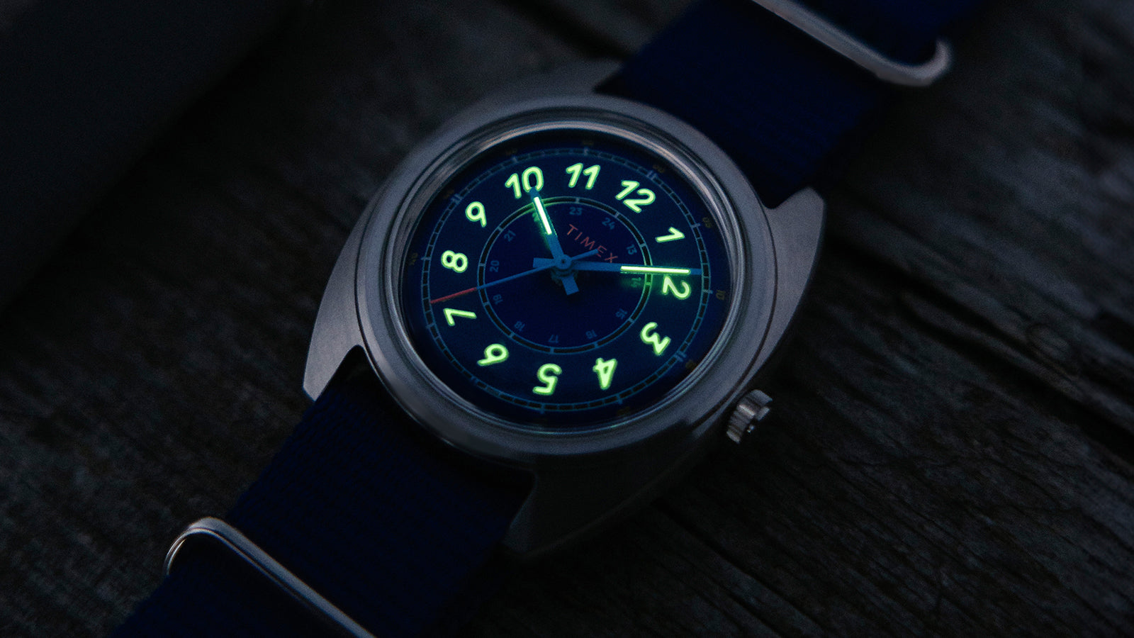 The Timex x Worn & Wound Whirlpool watch shown in low light, with the numerals and hands glowing bright green against the blue dial.