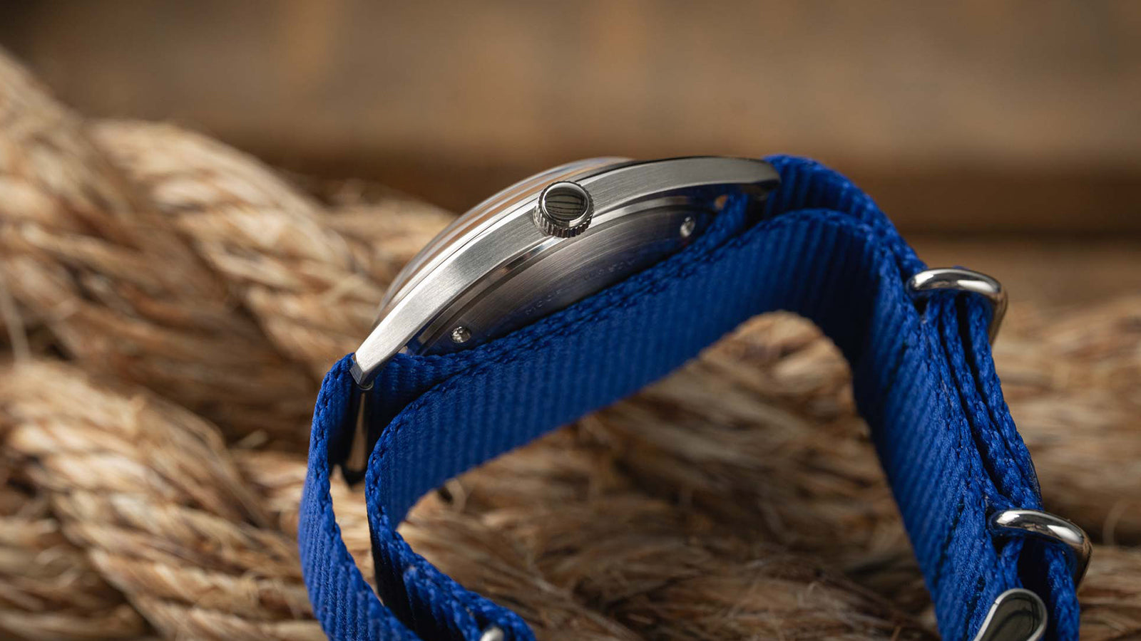 A side profile of the Timex x Worn & Wound Whirlpool watch, focusing on the brushed stainless-steel case, crown detail, and blue fabric strap wrapped over rope.