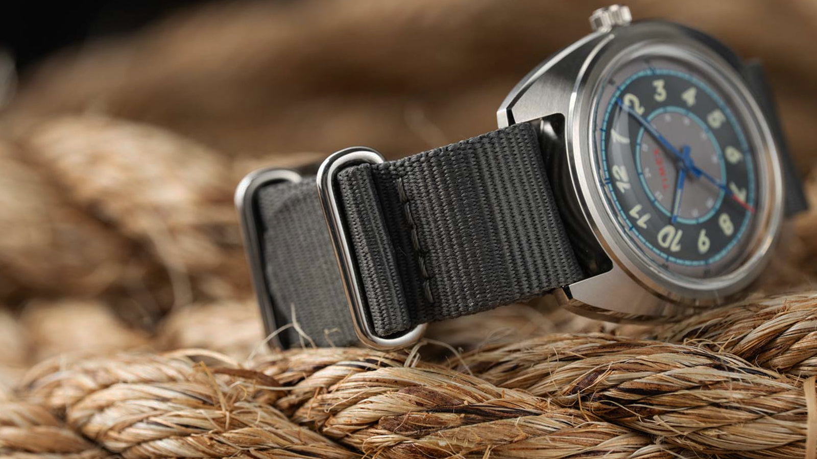 A side profile of the Timex x Worn & Wound Storm, highlighting the brushed stainless-steel case, domed crystal, crown, and gray fabric strap resting on rope.
