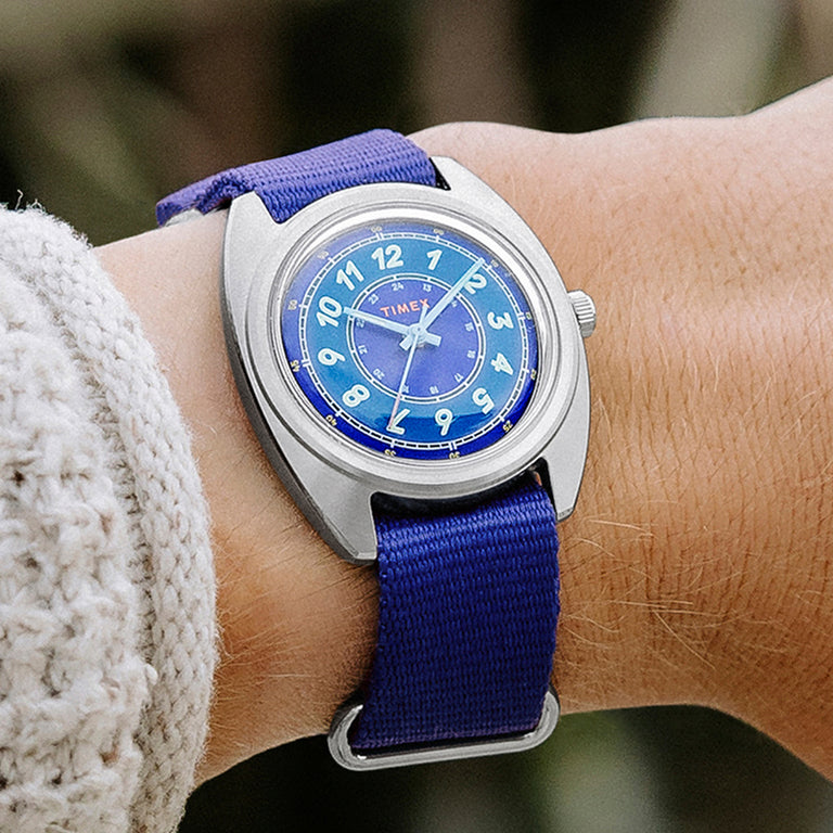 A close-up of a Timex x Worn & Wound wristwatch on a person’s wrist. The watch features a brushed silver-toned case, a vibrant blue dial with light blue numerals, and a matching blue fabric strap. The person is wearing a textured knit sweater, and the background is softly blurred.