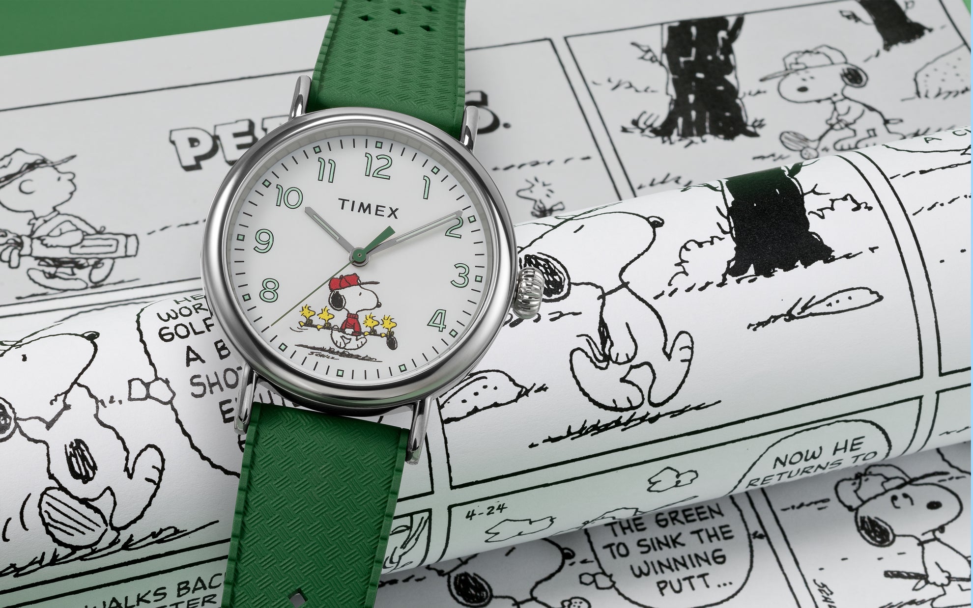 A Timex x Peanuts® Standard watch with a white dial and green strap rests on Peanuts comic strips. The dial features Snoopy dressed as a golfer in a red cap with three Woodstocks carrying golf clubs, blending sporty nostalgia with playful Peanuts charm.