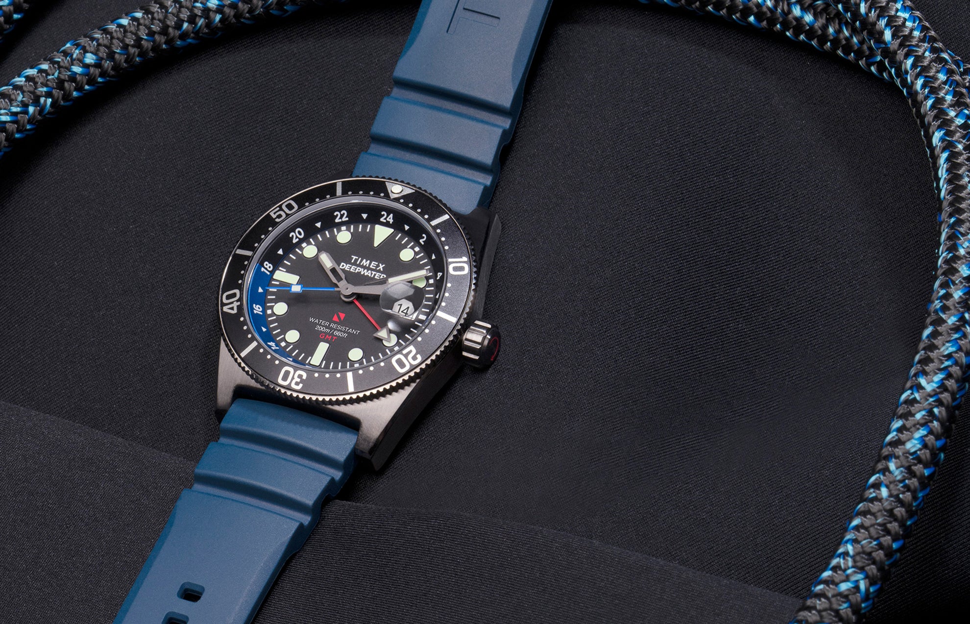 Timex Deepwater Reef 200 GMT dive watch with blue strap and black dial featuring a blue and black bezel, photographed against dark neoprene fabric and matching rope detailing.