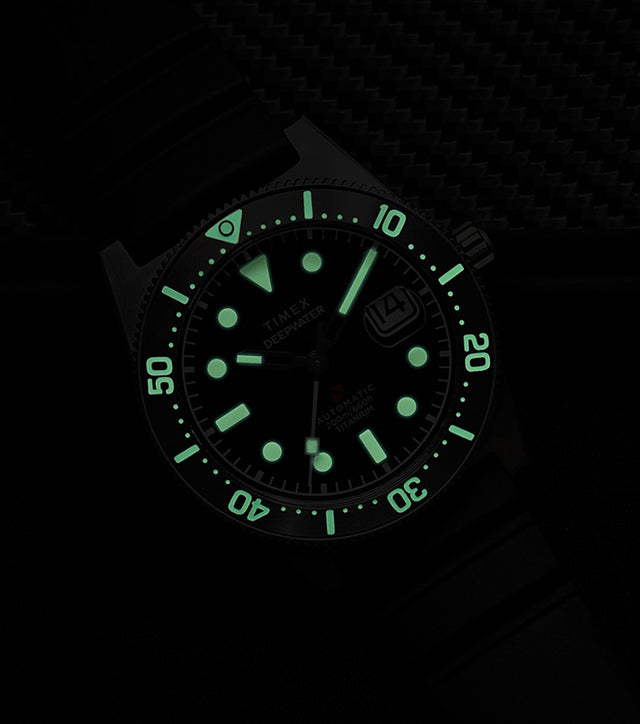 Timex Deepwater Reef 200 Titanium Automatic watch glowing in the dark, showing strong luminescence on the dial and bezel markers against a dark background.