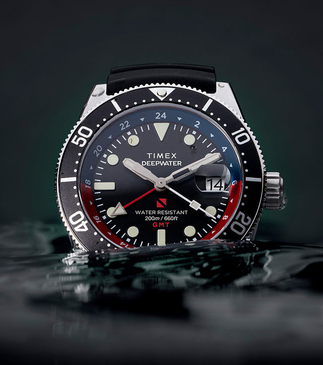 Timex Deepwater Reef 200 GMT dive watch with black dial, black strap, and red-blue bezel partially submerged in water, emphasizing its rugged design and 200-meter water resistance.