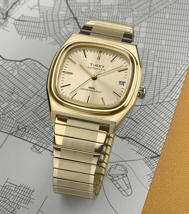 Timex® Automatic 1983 E Line Reissue watch in gold-tone stainless steel with an integrated bracelet and champagne dial. The watch is displayed on a detailed city map with a yellow textured accent in the background.