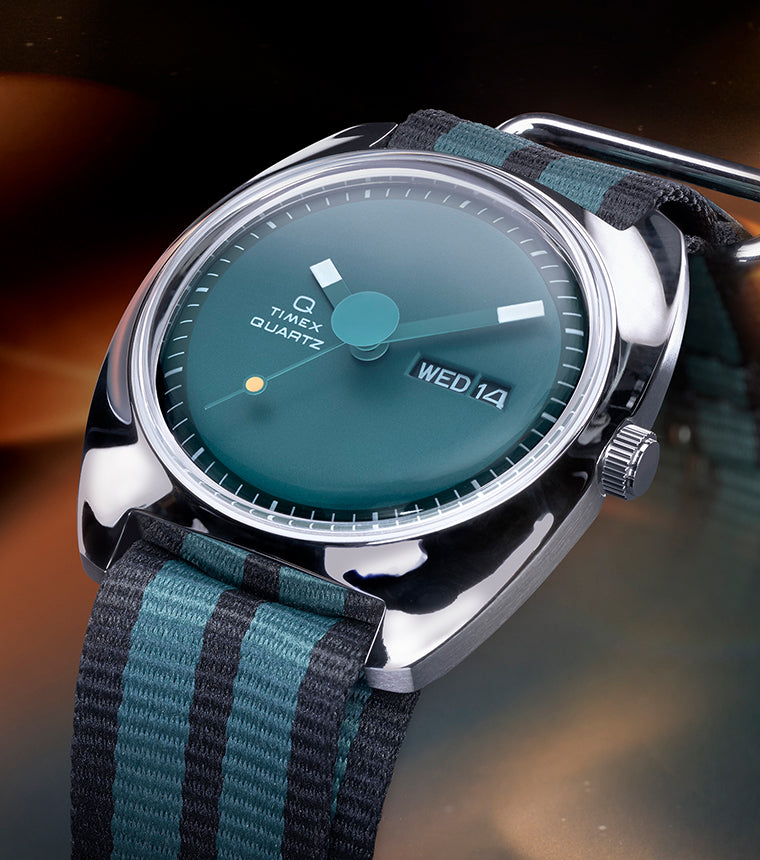 Angled view of the Q Timex® 1975 Enigma watch with a teal dial, polished silver-tone case, and black-and-green striped fabric strap, set against a glowing cosmic-inspired background.