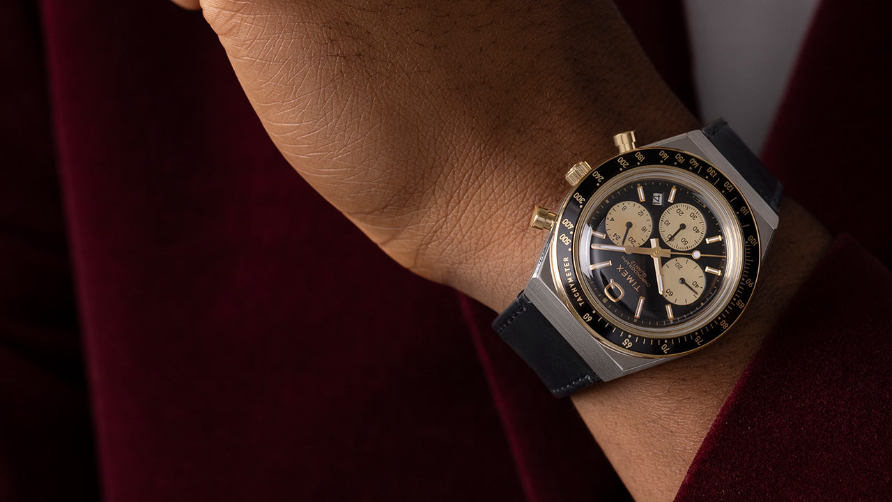 Q Timex Chronograph with black and gold dial worn on wrist, paired with a dark red jacket, showcasing a refined vintage-inspired look.