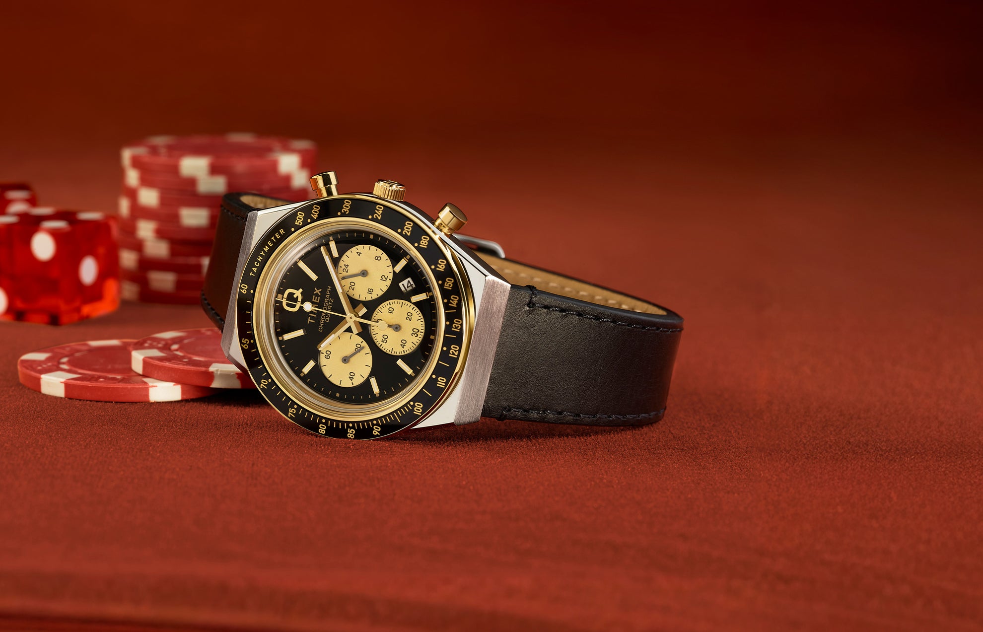 Q Timex Chronograph with a black dial and gold-tone sub-dials on a black leather strap, resting on a poker table with red chips and dice in the background.