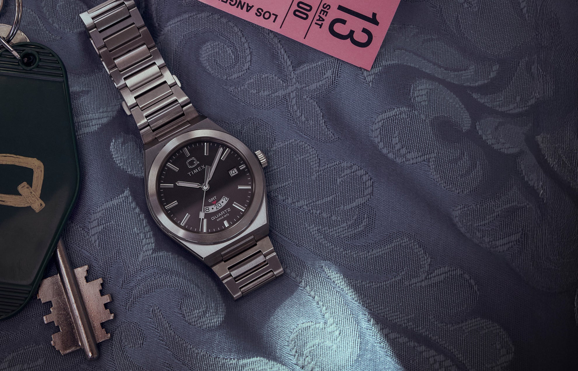 Q Timex® Continental GMT watch with a black dial, stainless-steel case, and bracelet displayed on a patterned blue fabric beside a vintage key and pink ticket stub.