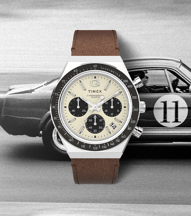Q Timex Chronograph watch with a cream dial, black sub-dials, and brown leather strap displayed over a vintage racing car background, evoking a classic motorsport aesthetic.