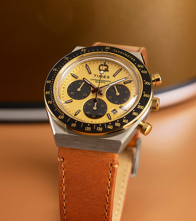 Q Timex Chronograph featuring a gold-tone dial, black sub-dials, and tan leather strap displayed upright on a warm gold background.