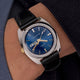 Q Timex® 1978 Day/Date watch worn on the wrist, showing the blue dial, gold-tone details, and black leather strap against a dark sleeve.