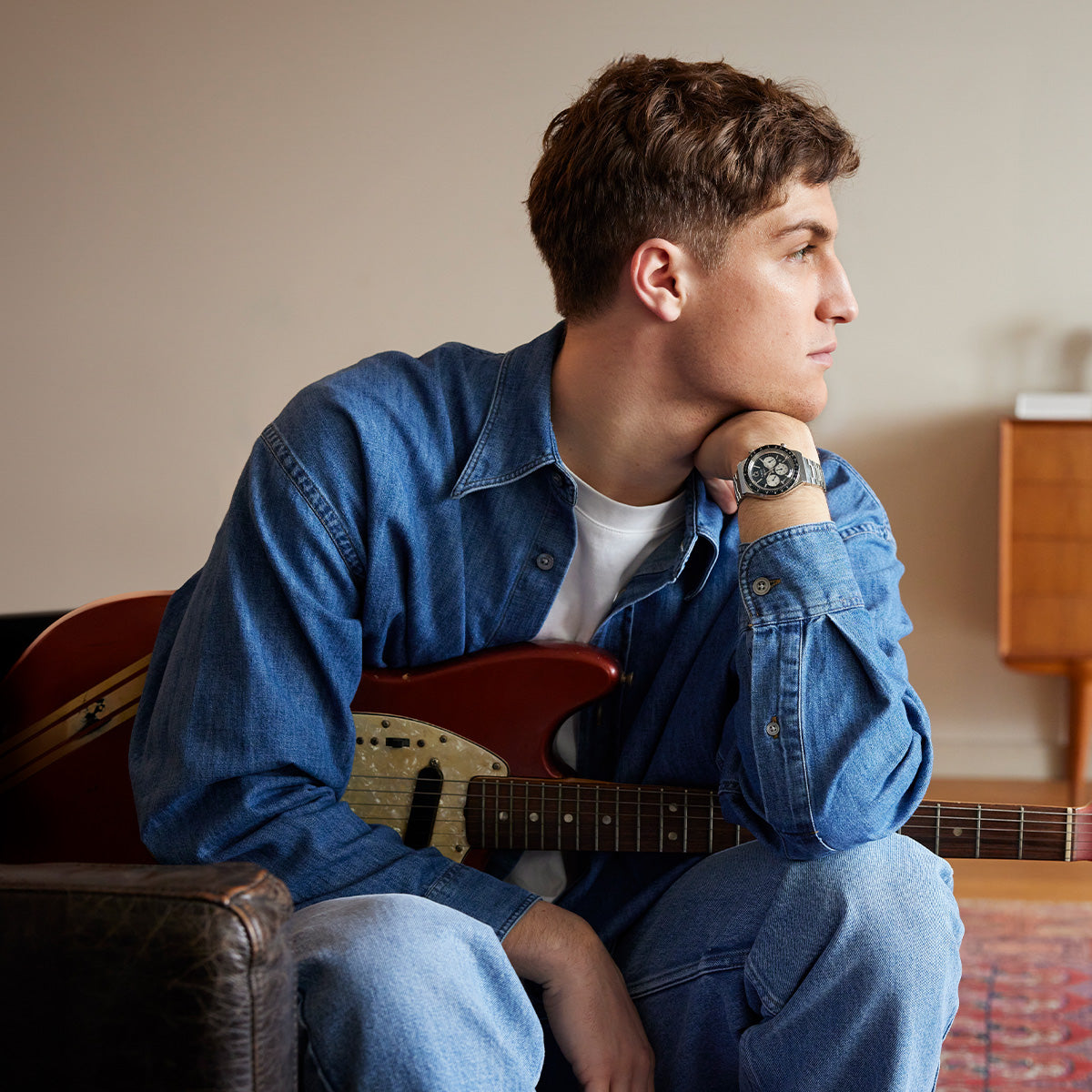 Person wearing a Q Timex Chronograph while holding a red electric guitar, dressed in denim shirt and jeans, in a relaxed home setting.