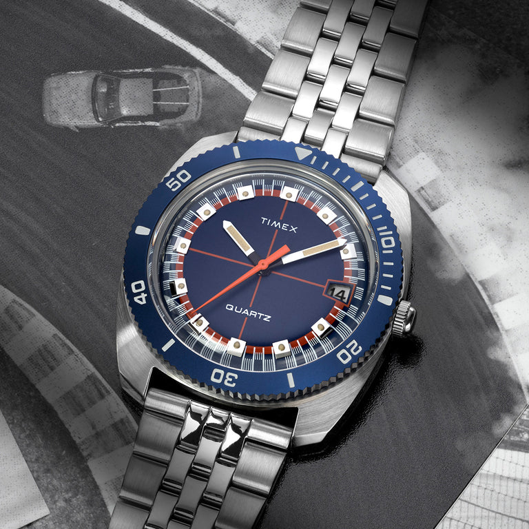Q Timex® 1971 Velocity watch with a blue dial and silver bracelet, placed over a black-and-white racing track photo, emphasizing its vintage motorsport inspiration.
