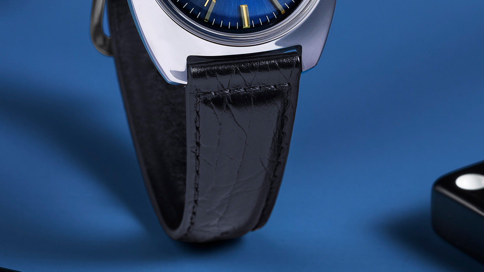 Detailed view of the Q Timex® 1978 Day/Date’s black leather strap with visible stitching and subtle texture on a blue surface background.