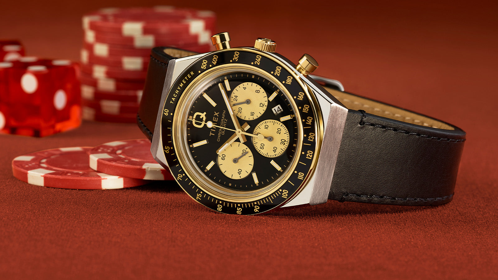Q Timex Chronograph with a black dial and gold-tone sub-dials on a black leather strap, resting on a poker table with red chips and dice in the background.