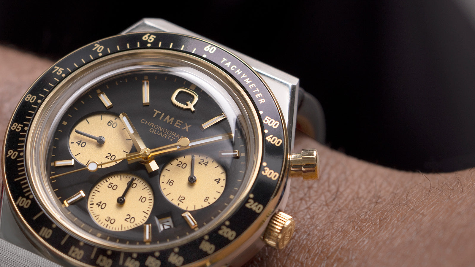 Close-up of the Q Timex Chronograph showing the black dial, gold sub-dials, and tachymeter bezel with polished gold accents and pushers.