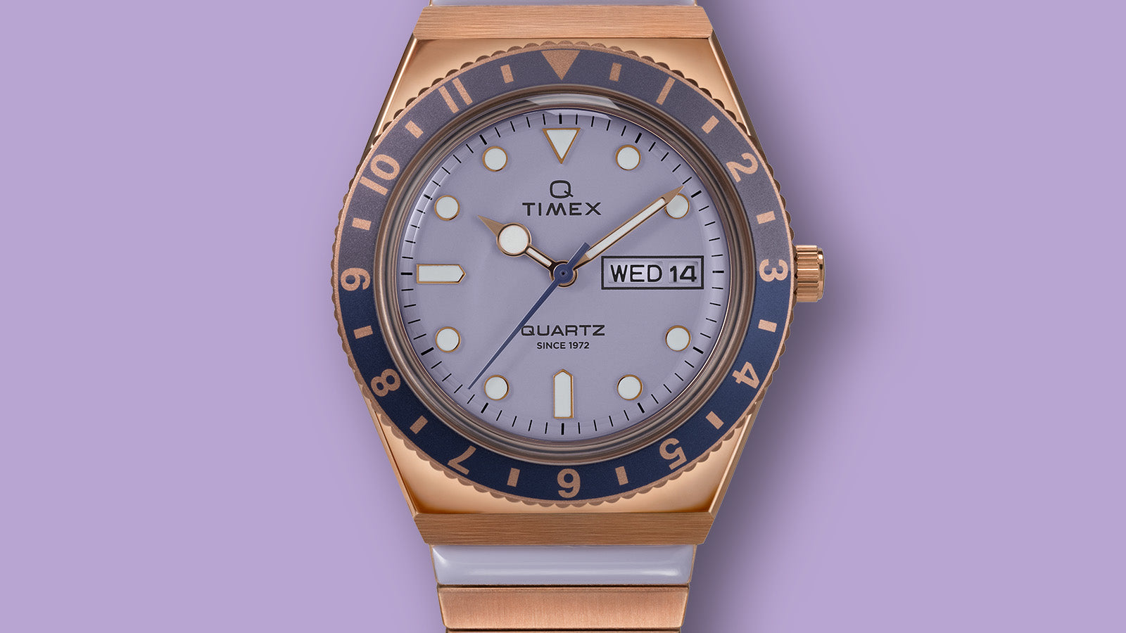 Front view of the Q Timex® watch showing the lilac dial, luminous hour markers, day-date window, and blue rotating bezel framed by a rose gold-tone case.