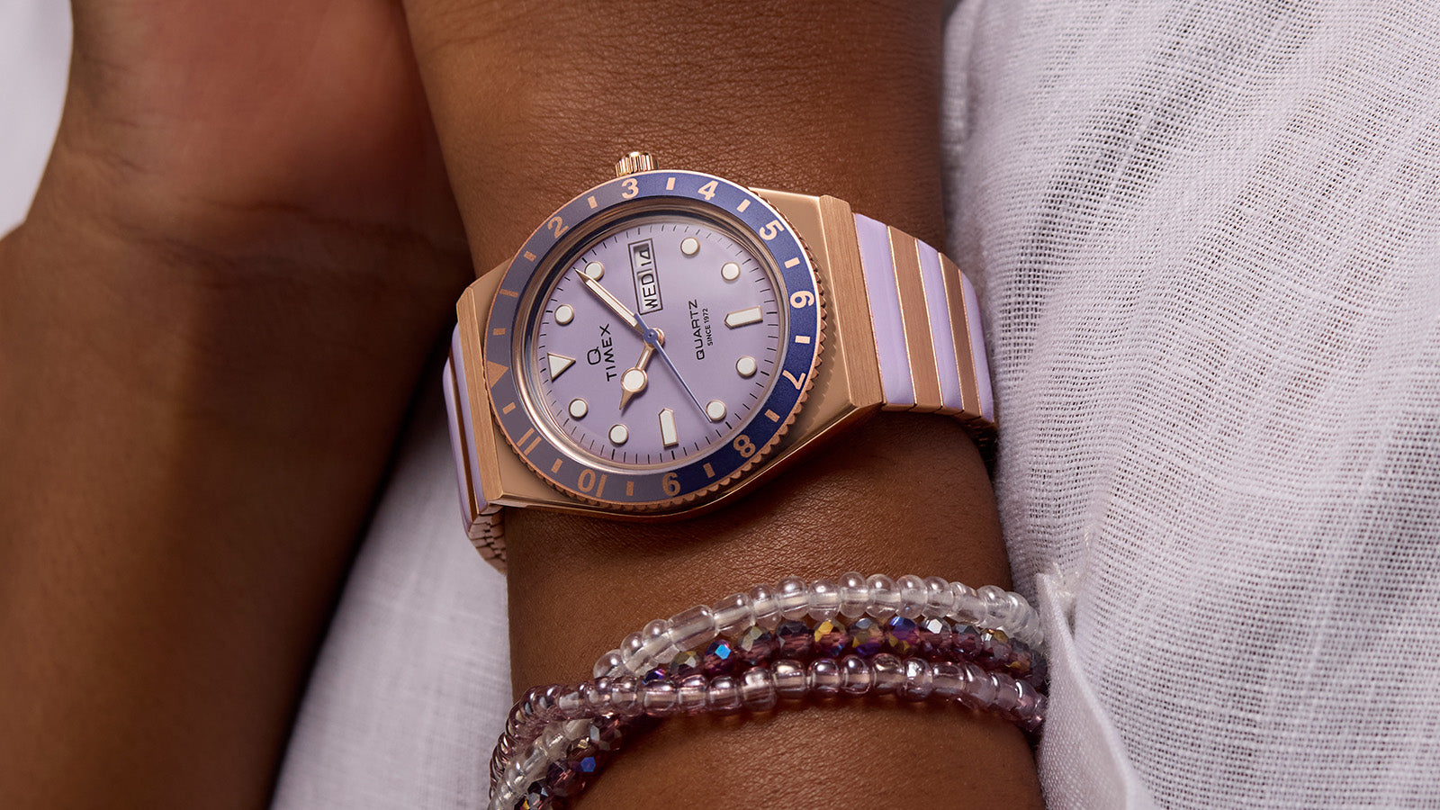 Q Timex® rose gold-tone watch with lilac dial and blue bezel worn on wrist, paired with beaded bracelets and a white textured blouse, creating a soft and stylish look.
